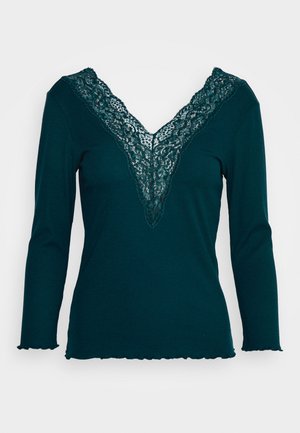 Donkergroenblauw gebreid top met lange mouwen, diepe V-hals afgezet met bijpassende kant en subtiele ruffles aan zoom en manchetten.