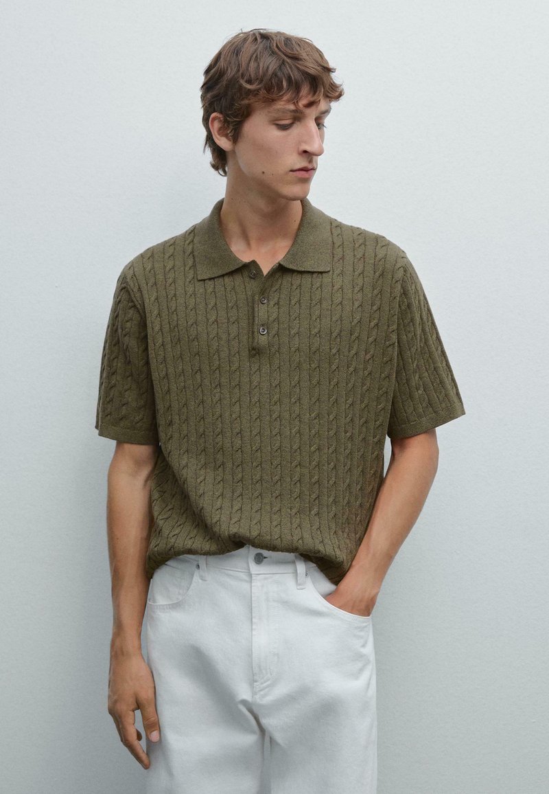Un giovane uomo si trova contro uno sfondo minimale, indossando un polo verde oliva con texture e pantaloni bianchi, guardando pensieroso di lato.
