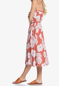 Robe midi corail avec un motif floral blanc, bretelles à nouer ajustables, taille cintrée et tissu léger. Associée à des sandales plates marron.