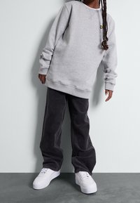 Szary sweatshirt z prążkowanymi mankietami, w połączeniu z czarnymi spodniami z sztruksu. Białe tenisówki z teksturowaną podeszwą. Małe żółte logo na klatce piersiowej.