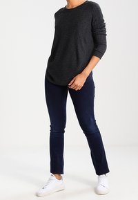 Person trägt einen dunkelgrauen Pullover mit langen Ärmeln, dunkelblaue enge Jeans und weiße Turnschuhe und steht mit einer Hand in der Nähe der Tasche.