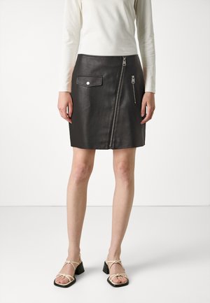 Kvinde iført en sort læder mininederdel med lynlåse foran og lomme, kombineret med en hvid bluse med lange ærmer og beige hælede sandaler.