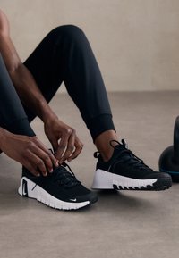 Scarpe da ginnastica nere con tomaia texturizzata, intersuola bianca e suola nera. Le stringhe vengono annodate da mani. Design semplice e lineare.