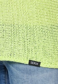 Neon green knitted top with an open weave. Tag on the lower edge features black background and white text, paired with light blue denim jeans.