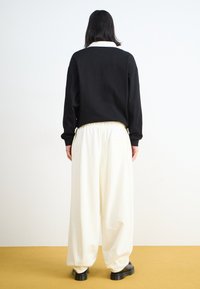 Black long-sleeve top with white collar, paired with loose white trousers, set against a plain background with a yellow floor.