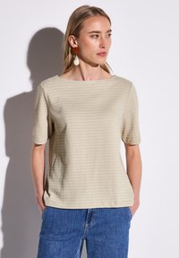 Beige texturovaný top s krátkým rukávem a širokým výstřihem, kombinovaný s modrými džínovými kalhotami. Látka má jemný geometrický vzor.