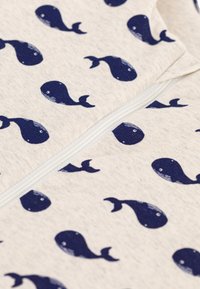 Raštuotas užtraukiamas drabužis, pagamintas iš šviesiai smėlio spalvos audinio pagrindo su tamsiai mėlynais banginių motyvais. Minkšta tekstūra ir subtilūs siūlių akcentai.