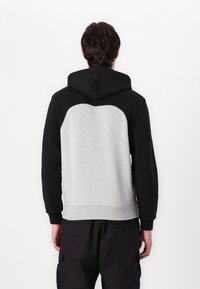 Sudadera con capucha que presenta un cuerpo gris y mangas negras. Textura suave con un corte recto y puños acanalados, la parte trasera muestra un diseño en bloques de color.