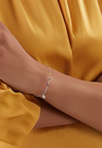 Pulsera colorida de cuentas con una cadena de plata y un pequeño charm plano, llevada en una muñeca, sobre una prenda de satén amarilla.