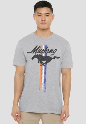 Junger Mann trägt ein graues T-Shirt mit schwarzem rennendem Pferd und dem Schriftzug "Mustang" über vertikalen Streifen in Orange, Weiß und Blau.