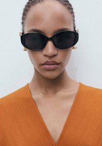 Black oval sunglasses with thick frames. Model wears an orange v-neck top. Smooth texture; minimal detailing. Neutral background.