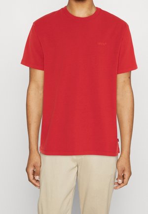 Mann trägt ein schlichtes rotes T-Shirt mit kurzen Ärmeln und einem dezenten Logo auf der Brust sowie beigefarbene Hose, steht vor einem hellen Hintergrund.