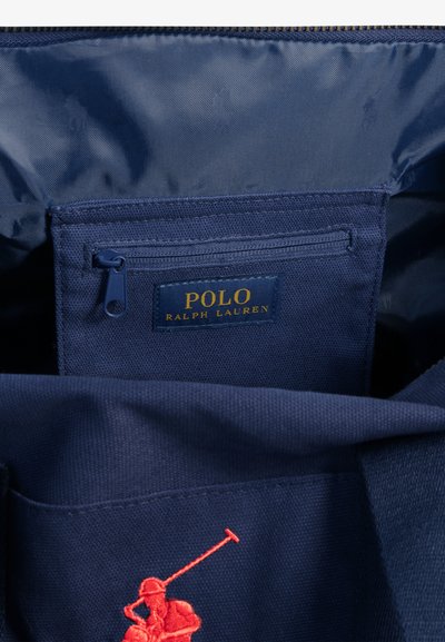 Interior de bolsa de lona azul marino con bolsillo con cremallera; presenta una etiqueta con el logo en oro y un diseño bordado en rojo de un jugador de polo. Textura suave y forma estructurada.