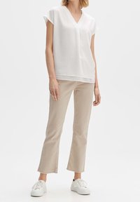 Weiße V-Ausschnitt-Bluse mit kurzen Ärmeln, kombiniert mit beigen, knöchellangen Hosen und weißen Sneakers, und damit ein lässiges Outfit kreierend.