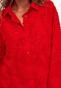 Chemise rouge à manches longues avec un tissu texturé et effiloché. Les détails notables incluent un col classique et une apparence translucide.