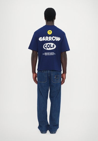 Tricou blue navy cu mânecă scurtă, cu "BARROW GOLF" imprimat în alb pe spate, asortat cu o pereche de blugi albaștri largi.