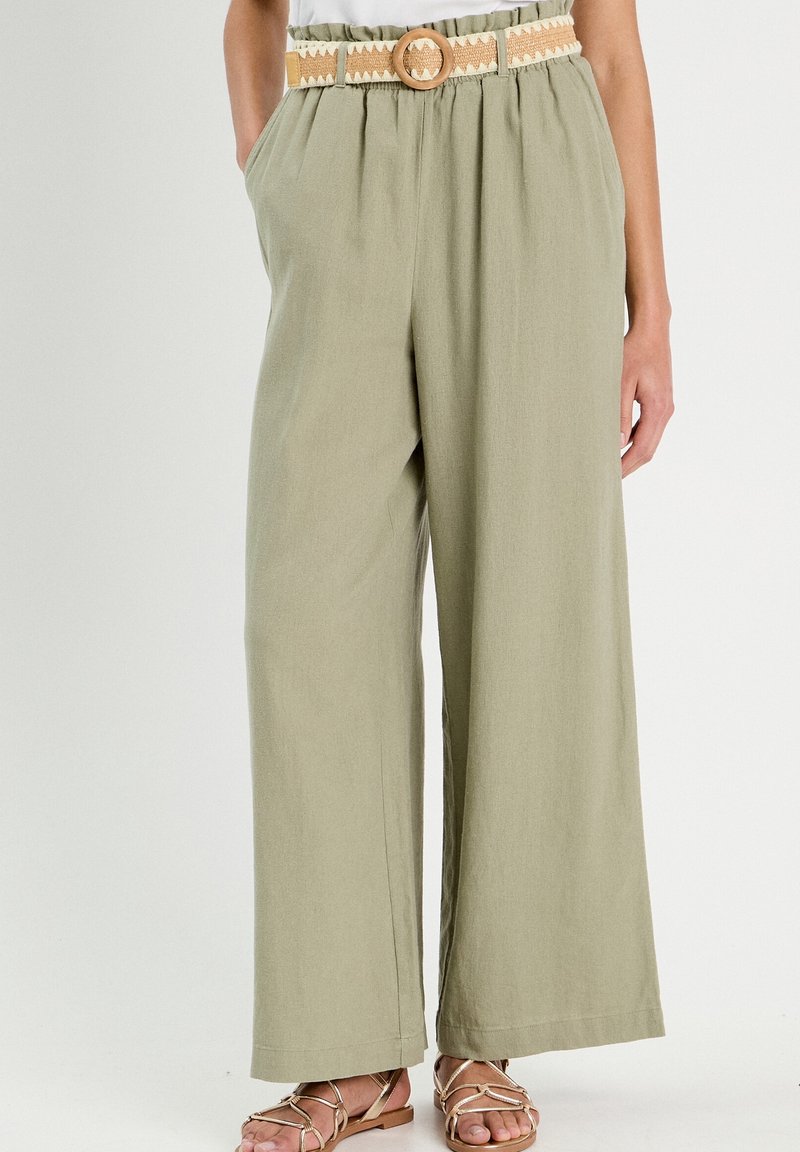Pantalon large couleur olive clair avec une ceinture beige tissée et une boucle ronde, porté avec un haut blanc et des sandales plates métalliques à brides.