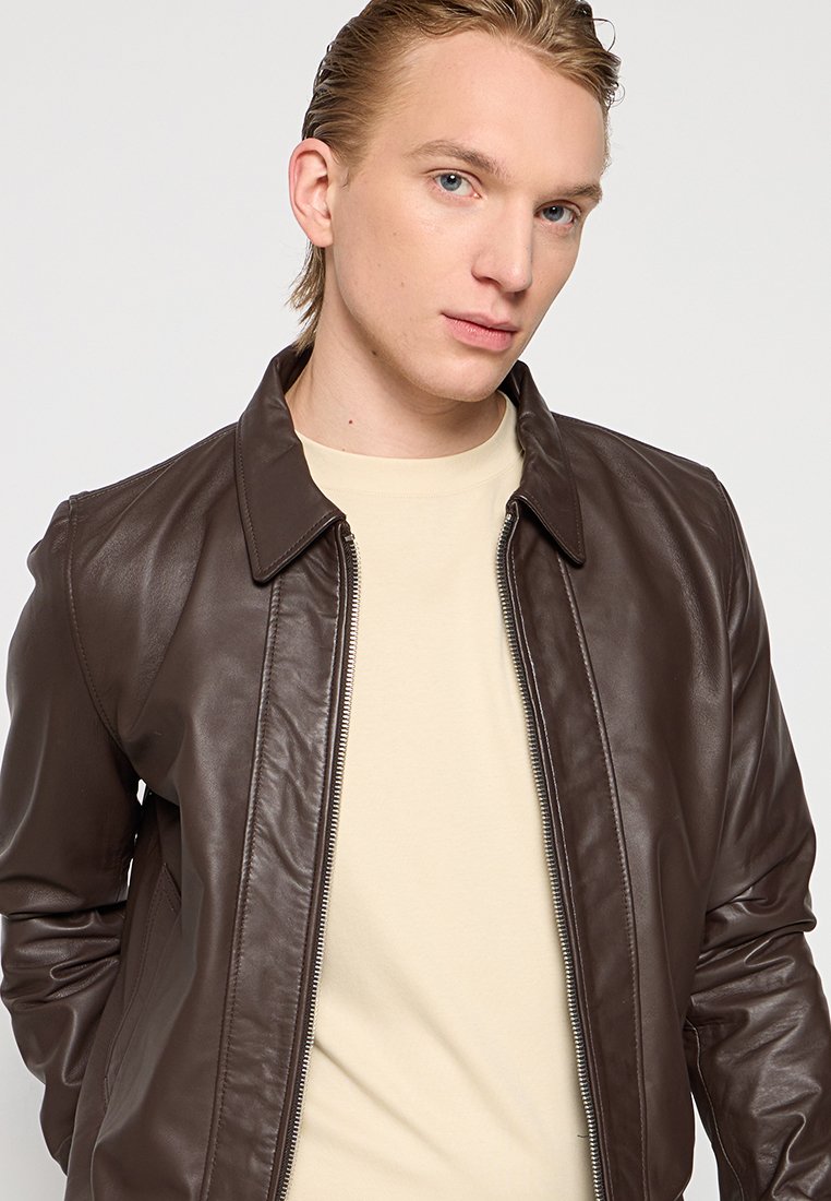 Veste en cuir marron avec fermeture éclair et col pointu. Portée sur une chemise beige, avec des manches longues et une texture lisse.