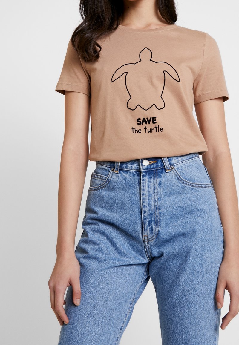 Personne portant un t-shirt beige avec un contour de tortue noir et le texte "SAVE the turtle,", associé à un jean bleu clair taille haute.