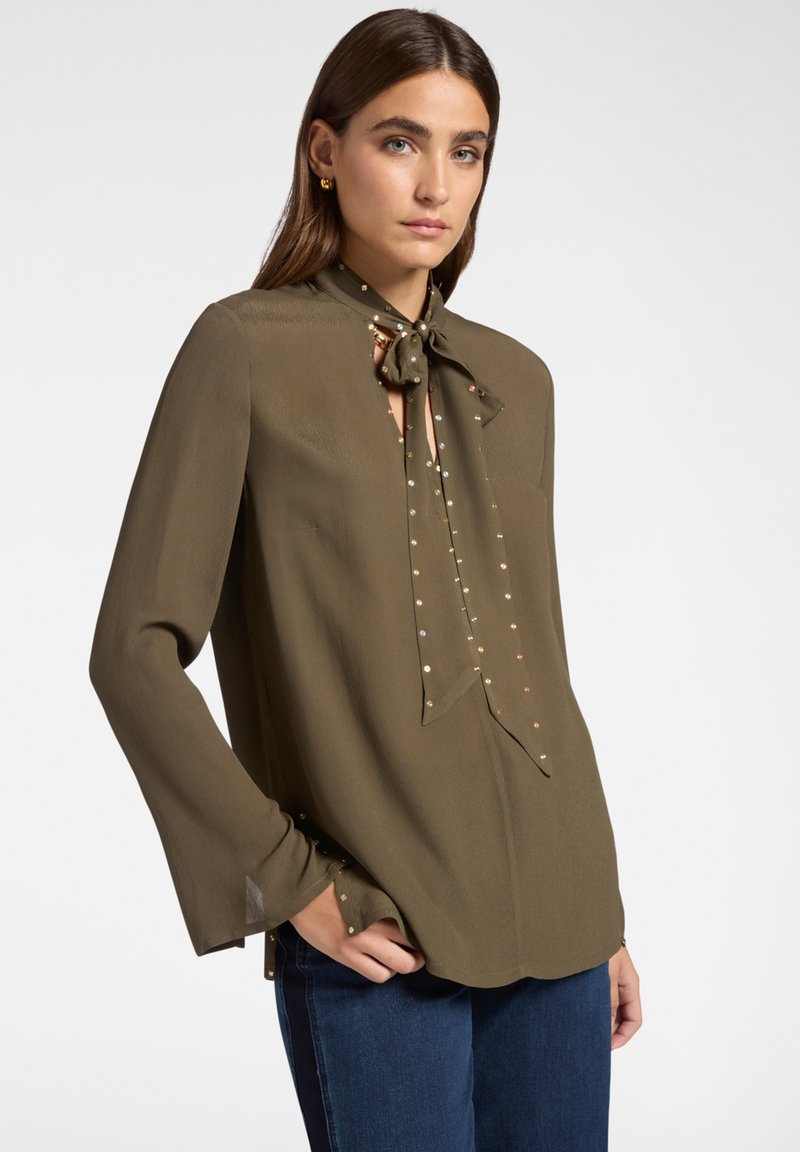 Donna che indossa una camicia a maniche lunghe verde oliva con collo a cravatta e piccoli ornamenti lucidi, abbinata a jeans blu, in piedi con una mano in tasca.