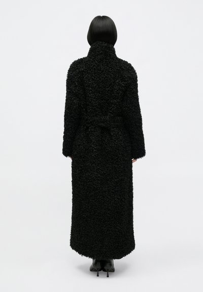 Casaco preto longo feito de material texturizado e peludo. Apresenta um colarinho alto, cintura cingida e cai até à metade da panturrilha. Salto alto preto simples.