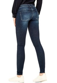 Persona con jeans skinny de color azul oscuro, camiseta de manga larga negra y zapatillas blancas de pie, de espaldas al espectador.