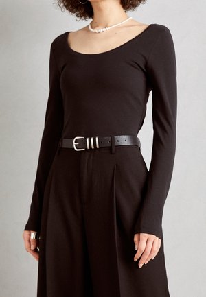 Vrouw die een zwart, lang mouws shirt met een ronde hals draagt, zwarte hooggetailleerde broek met riem, parelketting en zilveren ring aan haar linkerhand.