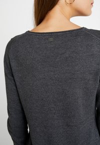 Pull gris foncé en tissu texturé, encolure ronde et petite étiquette logo au dos. Manches longues avec une coupe ajustée.
