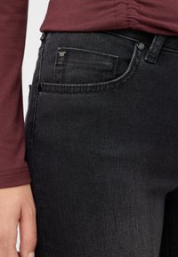 Czarne jeansy z denimu z widokiem zbliżeniowym, pokazującym przednią kieszeń, teksturowany materiał i metalowe akcenty. Towarzysząca bluzka jest burgundowa.