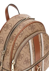 Zaino marrone con motivo, dotato di due scomparti con zip, dettagli in pelle color tan, strisce bianche e hardware dorato. Design arrotondato con una maniglia superiore.