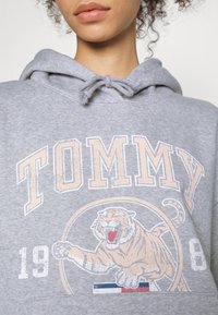 Harmaa huppari, jossa on kiristysnauhallinen kaula-aukko. Siinä on suuri "TOMMY"-painatus ja tiikerikuvio. Designissa on tekstuuriyksityiskohtia ja logo.