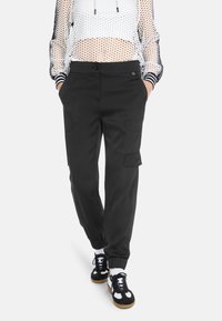 Schwarze Cargo-Hose mit Seitentaschen, elastischen Bündchen und glatter Textur, kombiniert mit einem weißen Mesh-Oberteil und schwarzen Sneakers.