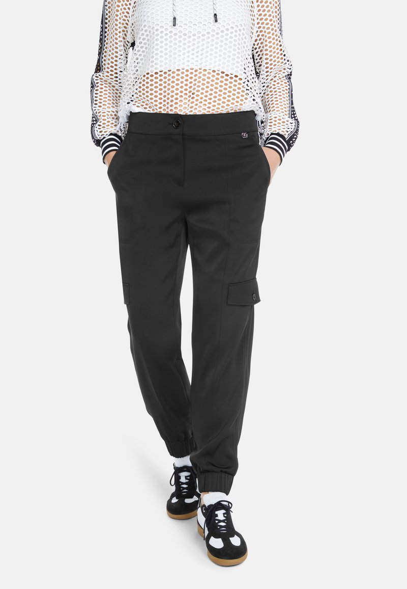 Schwarze Cargo-Hose mit Seitentaschen, elastischen Bündchen und glatter Textur, kombiniert mit einem weißen Mesh-Oberteil und schwarzen Sneakers.