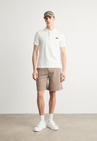 Biała koszulka polo z czarnym logo, beżowe szorty i białe sneakersy. Model nosi również beżową czapkę, stojąc na neutralnym tle.
