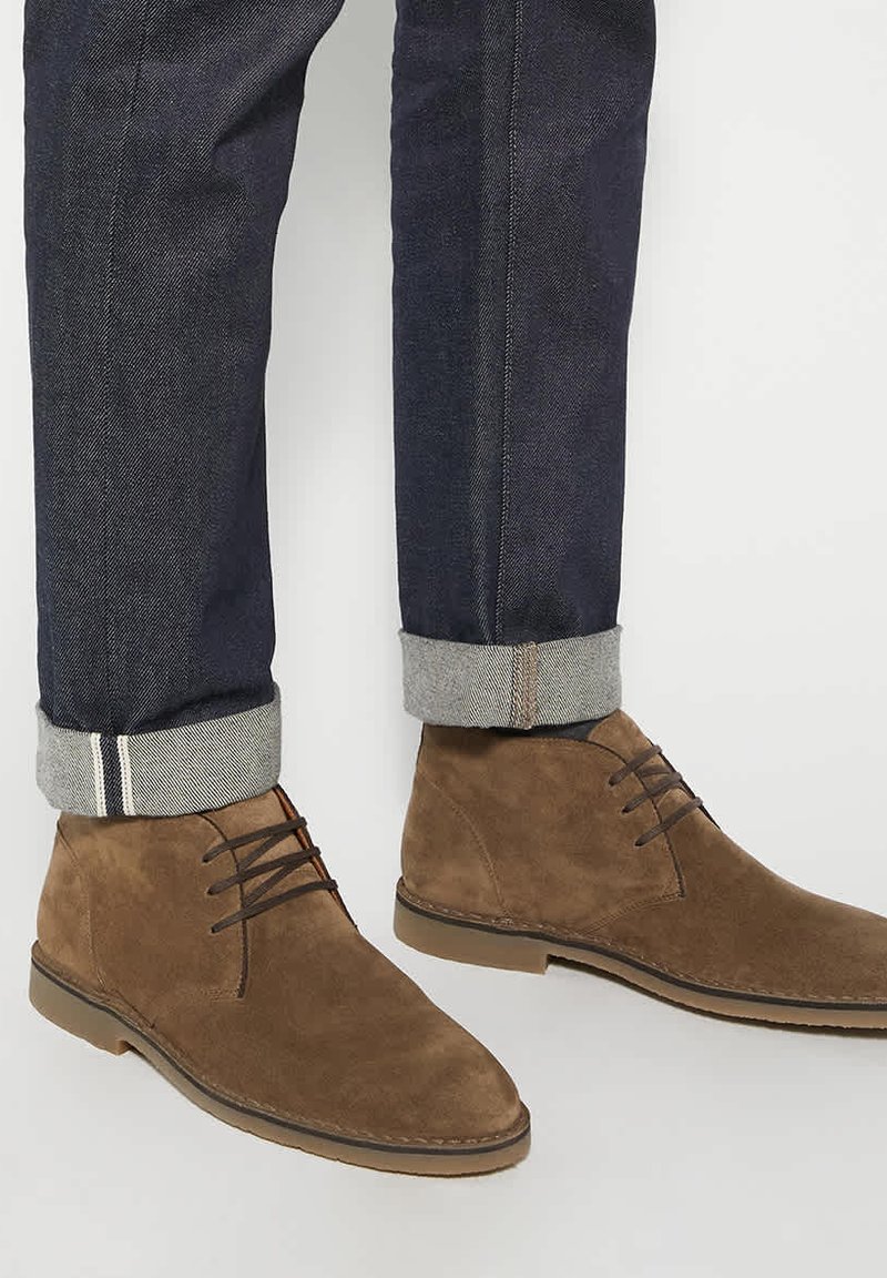 Bottines en daim marron à lacets portées avec un jean en denim foncé retroussé sur un fond blanc.