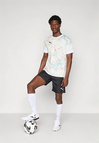 Mees valges mustriga särgis, mustades pükstes ja valgetes sokkides, kes seisab ühe jalaga mustvalgel jalgpallil ühevärvilise tausta ees.