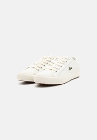 Sneakers bianchi in tela con una forma a bassa altezza, dotati di chiusura con lacci e suole in gomma testurizzata. Piccolo dettaglio del logo verde sul lato.