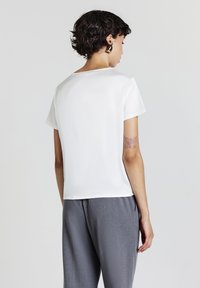 Donna con capelli scuri e corti, che indossa una maglietta bianca a maniche corte, pantaloni grigi, orecchini d'oro e un tatuaggio di farfalla sul braccio sinistro.