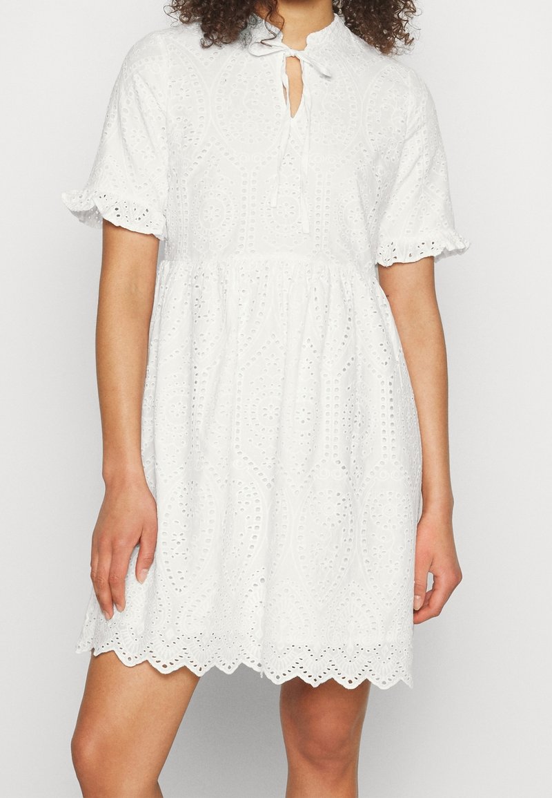 Robe blanche en dentelle à œillets avec manches courtes bouffantes, ourlet festonné et encolure goutte avec lien, portée par une personne debout.