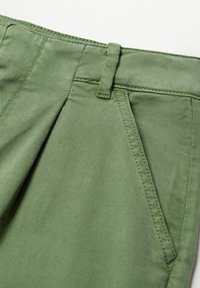 Pantalon en tissu vert en gros plan montrant la taille, la passants de ceinture, le pli et la poche latérale inclinée avec une couture détaillée.
