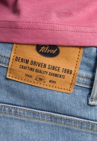 Un tessuto rosa con una superficie liscia sovrappone un cinturino in denim. Un'etichetta in pelle color sabbia con testo inciso reca la scritta "Petrol! Denim Driven Since 1989."
