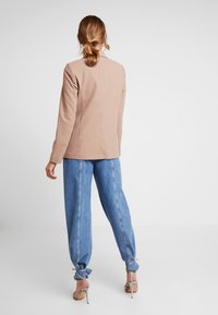 Blazer beige de diseño ajustado, mangas largas y líneas limpias, combinado con vaqueros anchos azul claro y sandalias metálicas con tacón.