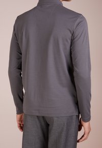 Koszula z długim rękawem w jednolitym odcieniu szarości, wykonana z lekkiego materiału, z wysokim kołnierzem i subtelnymi detalami szwów na plecach. Połączona z szarymi spodniami.