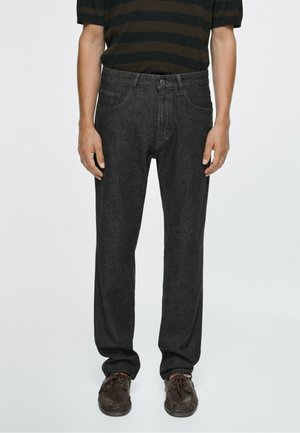 Homme portant un jean gris foncé droit, des chaussures en cuir marron et une chemise à manches courtes rayée noire et marron, debout avec les bras détendus.