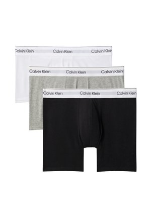 Homme musclé aux cheveux courts debout pieds nus sur un fond gris, portant un boxer Calvin Klein gris clair avec une taille blanche.