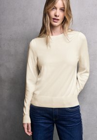 Maglione color crema con scollo rotondo e maniche lunghe, dotato di orlo e polsini a coste, abbinato a jeans blu. Texture liscia.