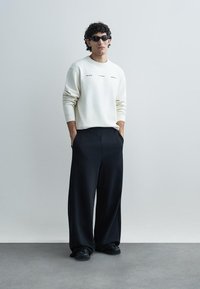 Biela mikina s potlačeným textom, kombinovaná s voľne strihanými čiernymi nohavicami. Doplnky zahŕňajú tmavé slnečné okuliare a čierne tenisky.