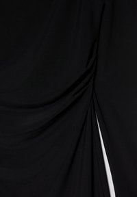 Fekete szövet, mely puha, sima tapintású, összegyűrt redőkkel és egy hasítékkal díszített, ami fokozza lágy, folyékony sziluettjét.