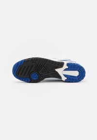 Semelle de chaussure de sport blanche avec une bande de roulement en caoutchouc noir et bleu, présentant des motifs variés et un dessin circulaire près de la plante du pied.
