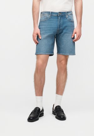 Mężczyzna w średnioblękitnych dżinsowych szortach, białych skarpetkach za kostkę, czarnych mokasynach i białej koszulce stojący na jednolitym tle.